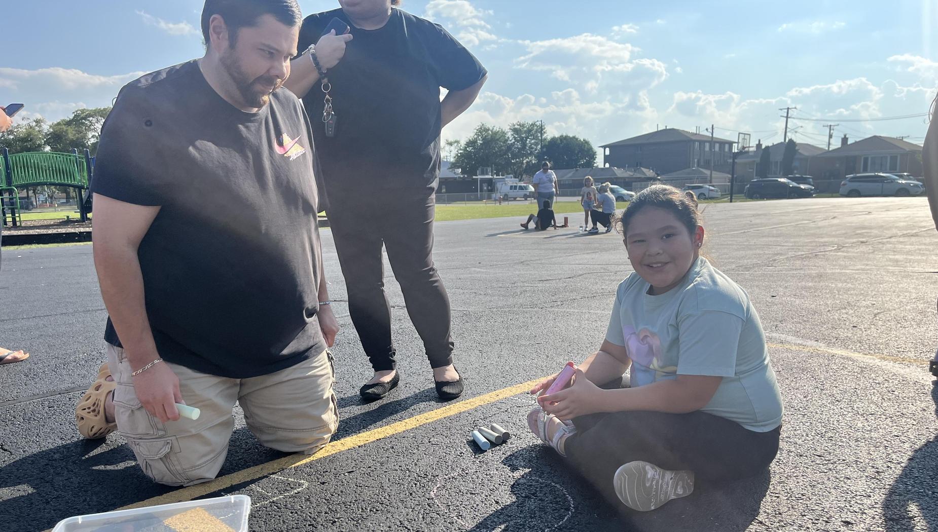A young girl sits on asphalt with chalk, while adults interact nearby in a sunny playground.