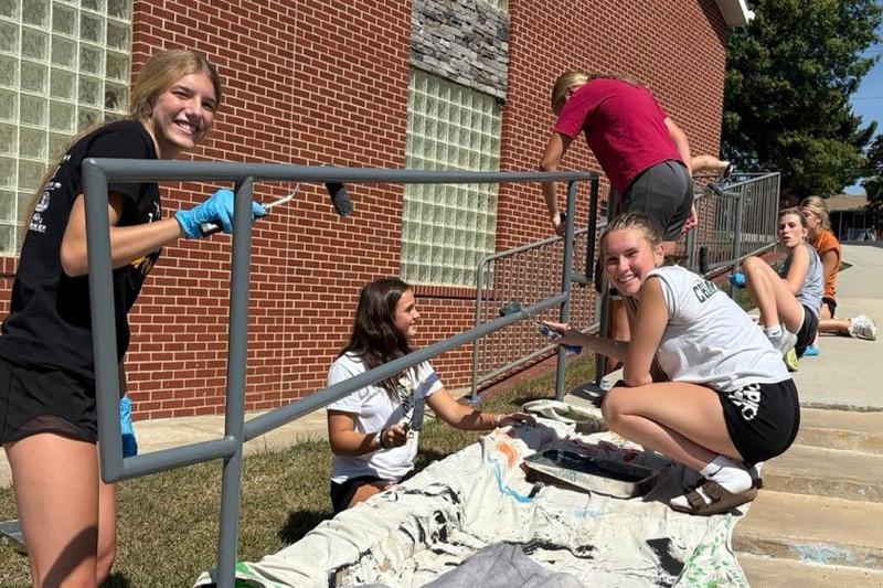 Liv Weishaar, Brooke Boss, and Kate Welsh paint a railing with their teammates