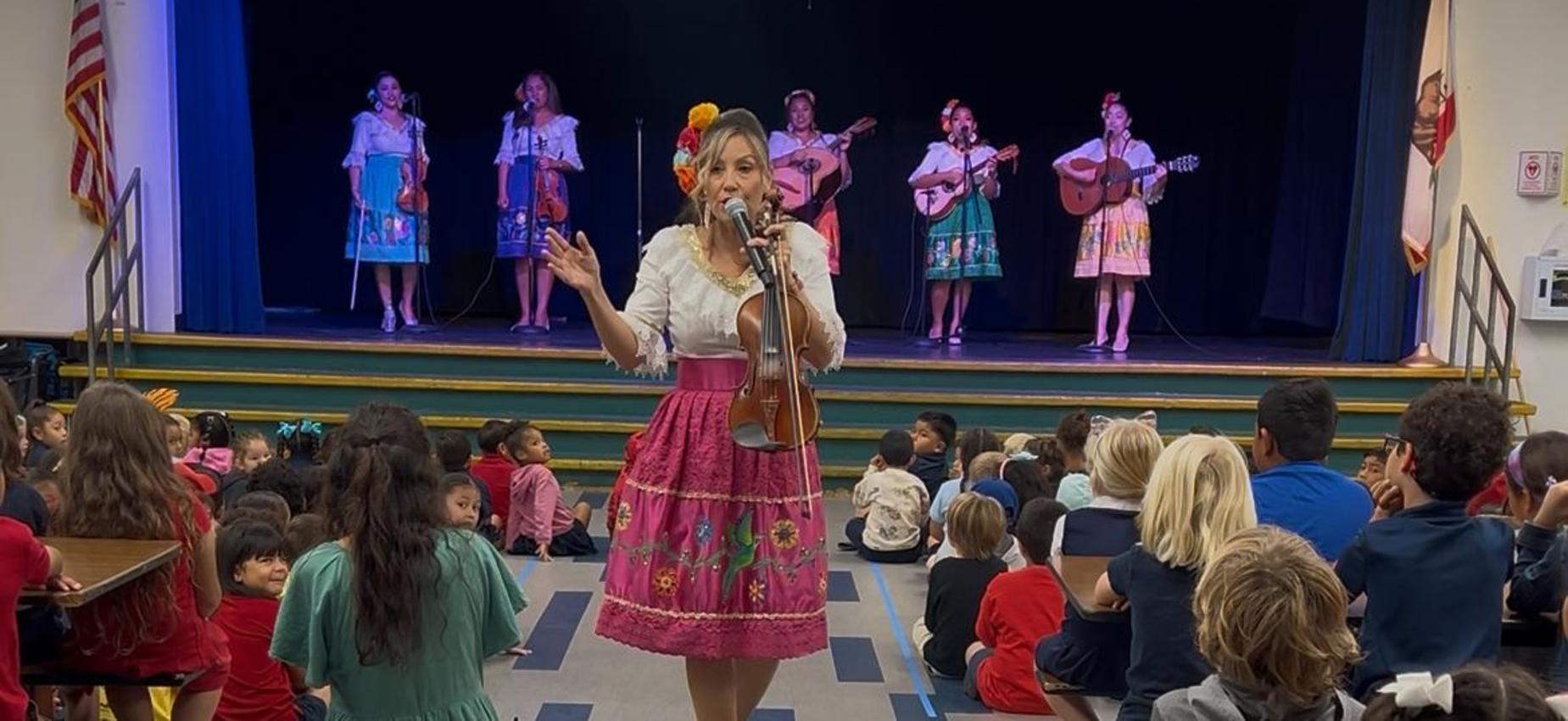 A woman in a colorful skirt plays the violin for an audience of children sitting on the floor.