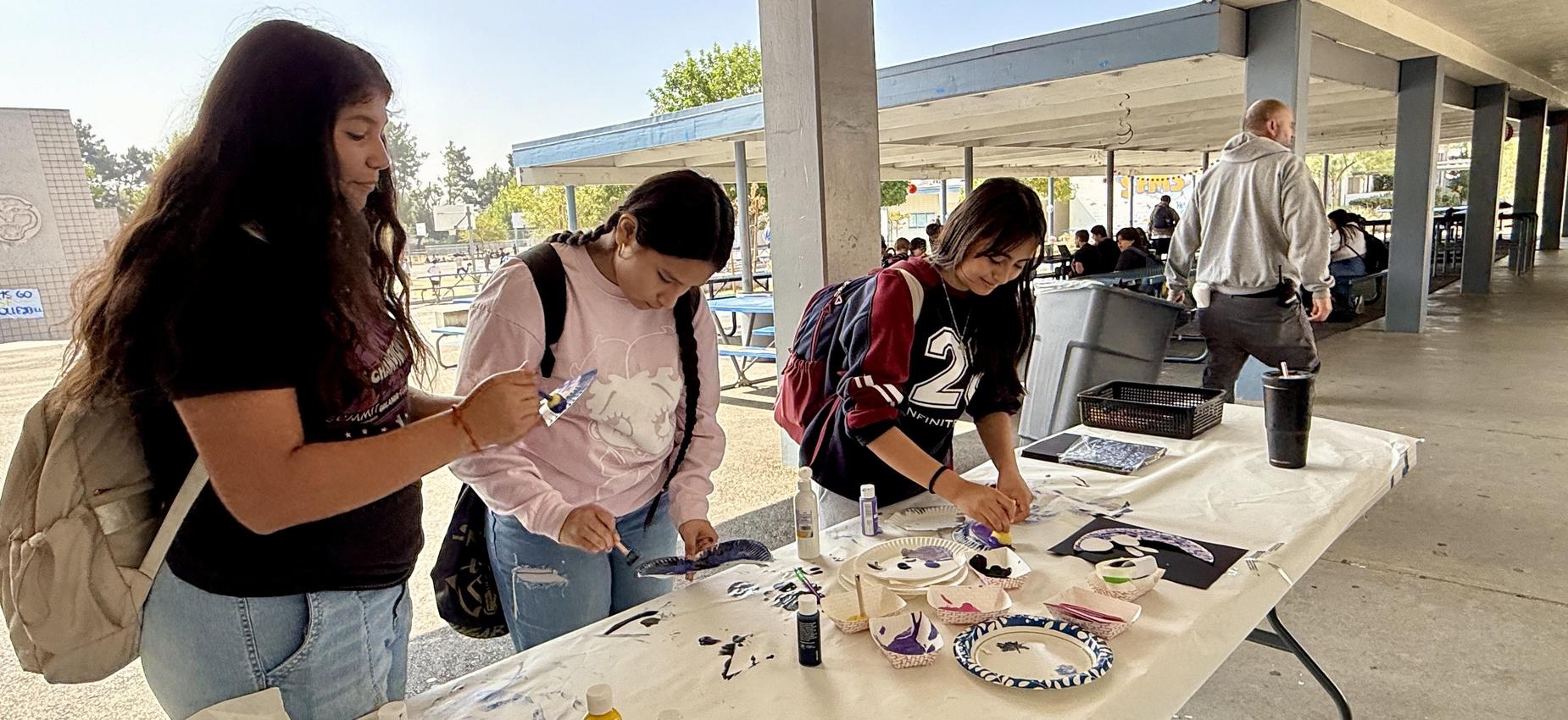Three students painting at a table outside, with various art supplies and classmates visible.