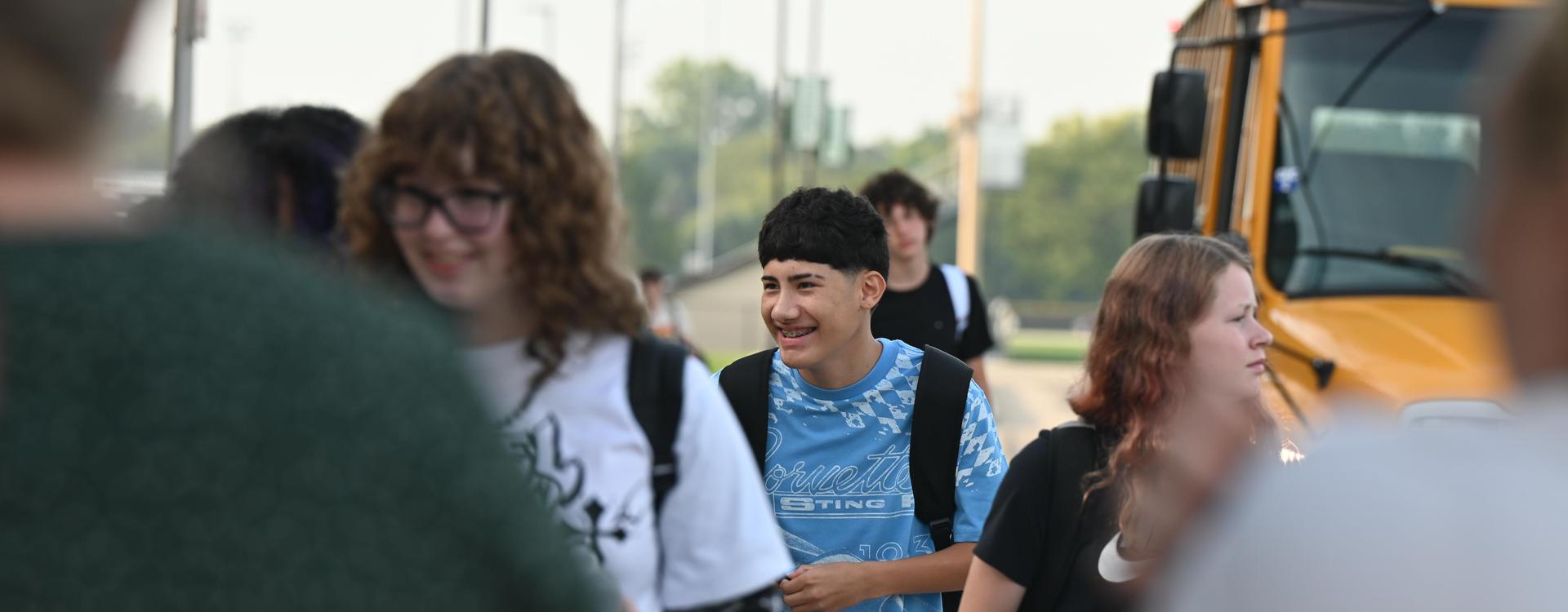 Students smiling and walking near a school bus in a busy outdoor setting.