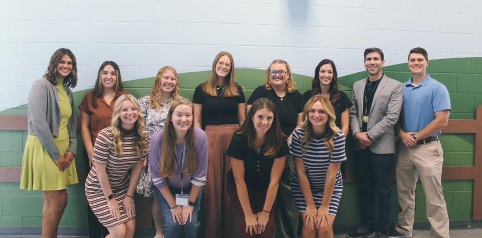 Group of people posing together in a classroom setting with a green background.