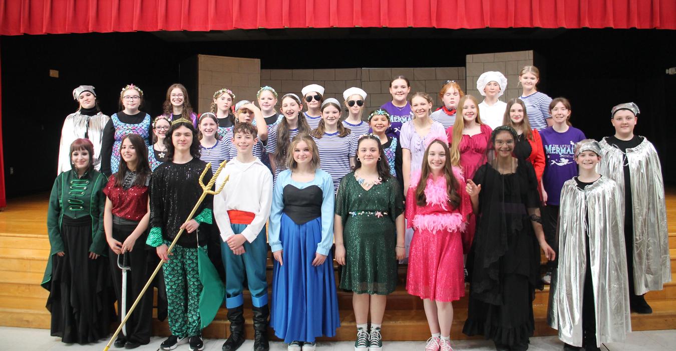 A large group of students in colorful costumes posing on stage with a red curtain backdrop.