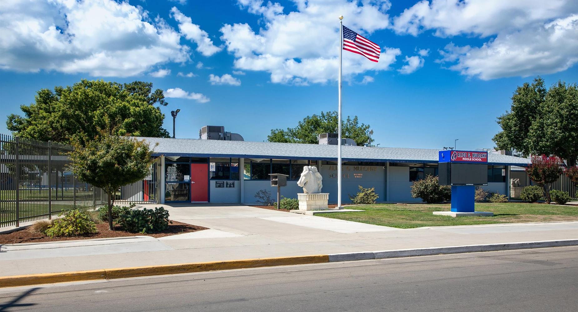 Mulcahy Middle School building with flag, Stallion statue, and landscaped area under a blue sky.