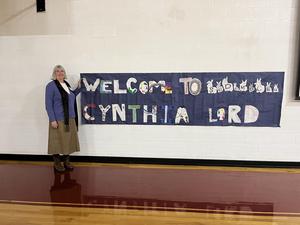 A woman stands next to a colorful welcome banner in a gymnasium.