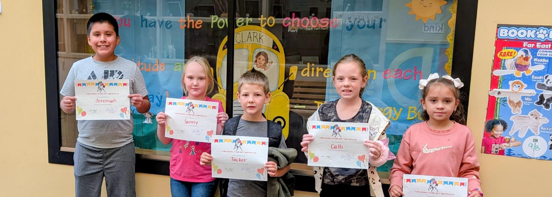 Group of six children holding achievement certificates in front of a colorful background.