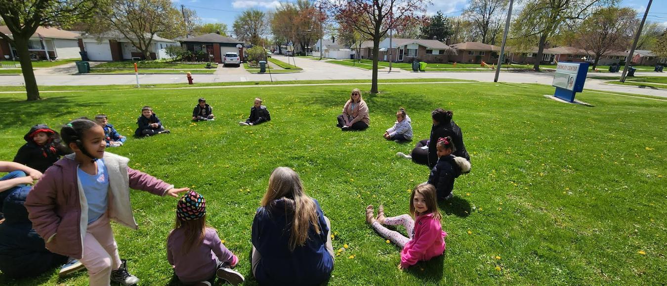 Children are outside sitting in a circle in the grass playing Duck, Duck, Goose.