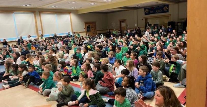 Large audience of children seated together in a hall, engaged and clapping.