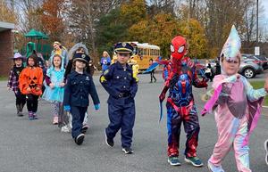 Children in costumes, including superheroes and a police officer, walk together during a parade.