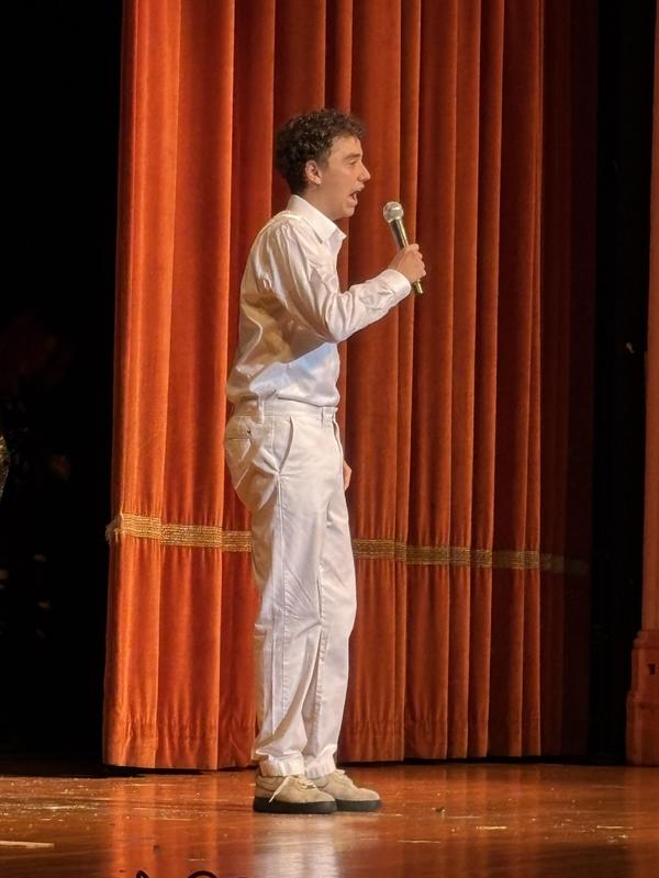 A performer dressed in white speaking into a microphone on stage.