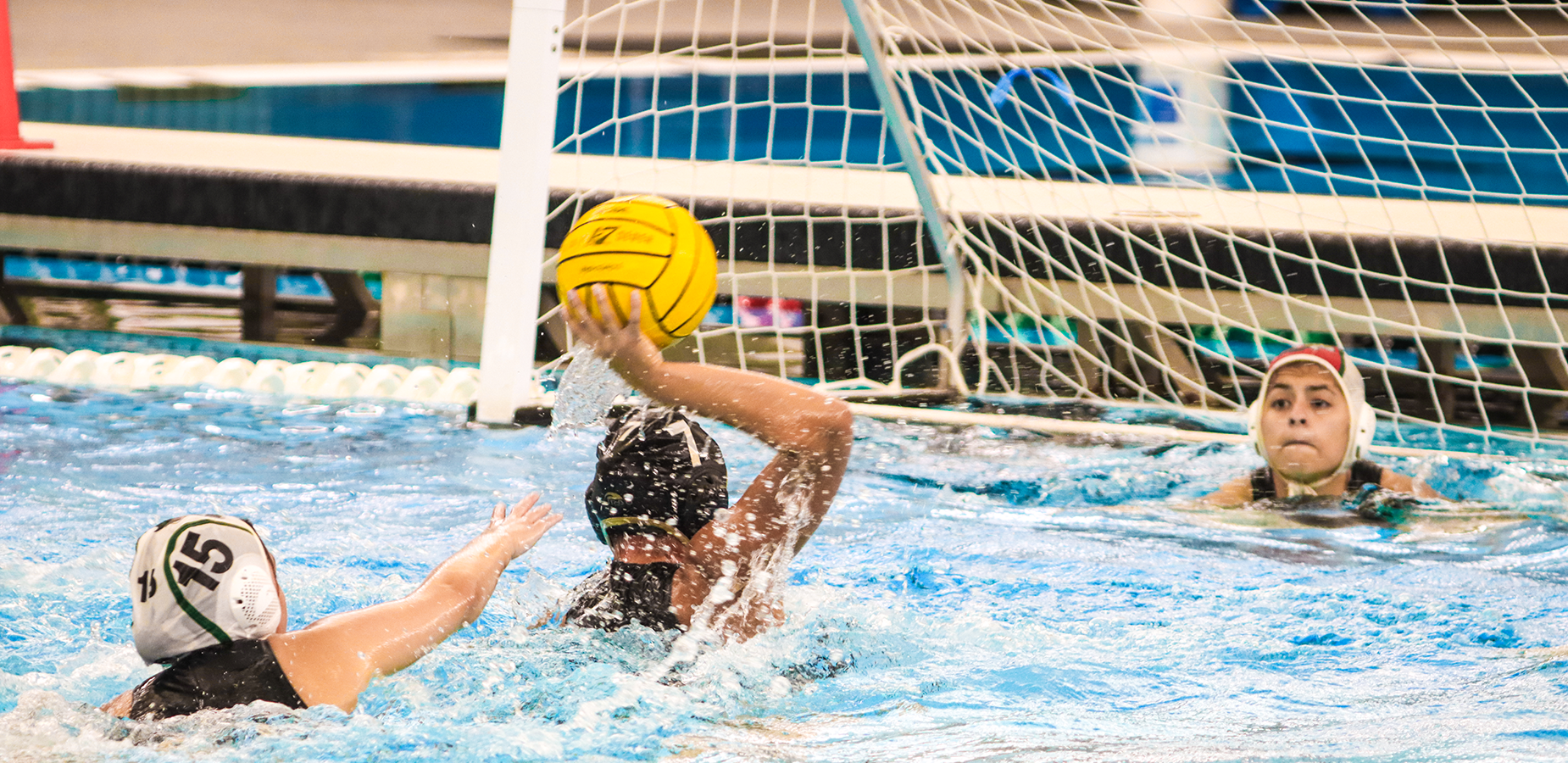 Water polo players in action, one shooting at the goal while a goalie defends.