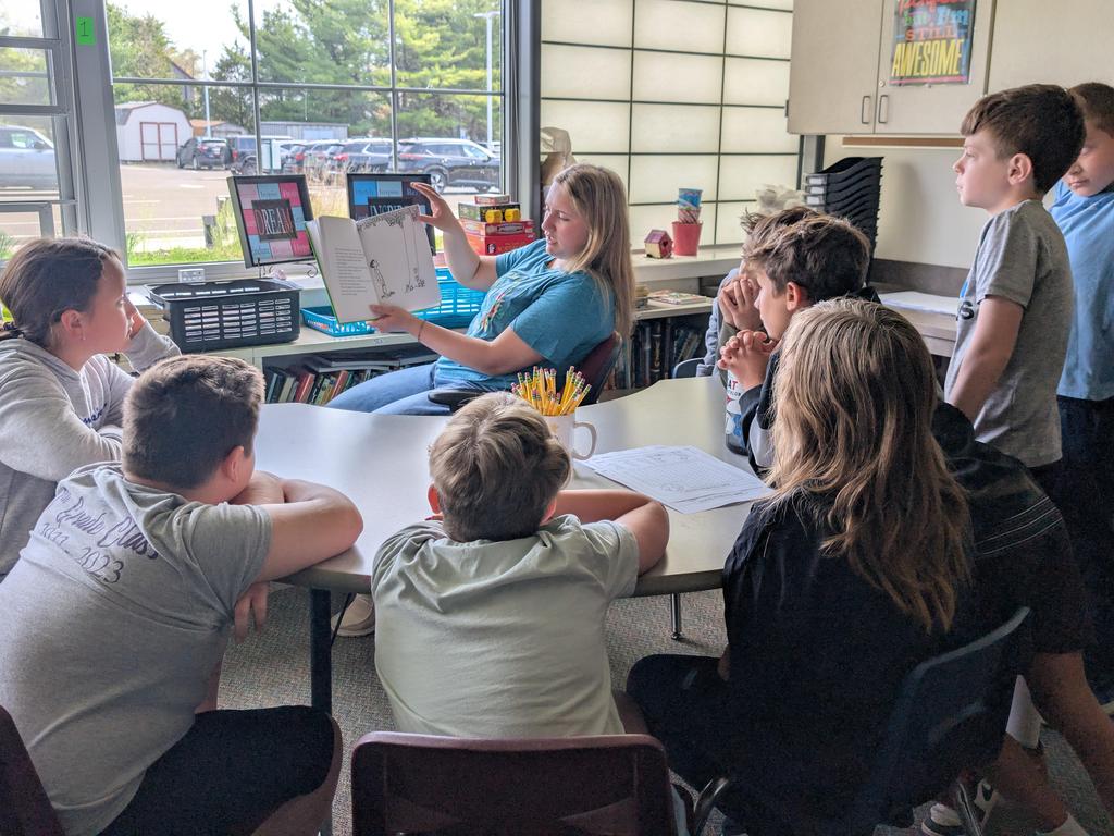 Students sitting at a table in a semi-circle and a teacher holding up a book to show them.