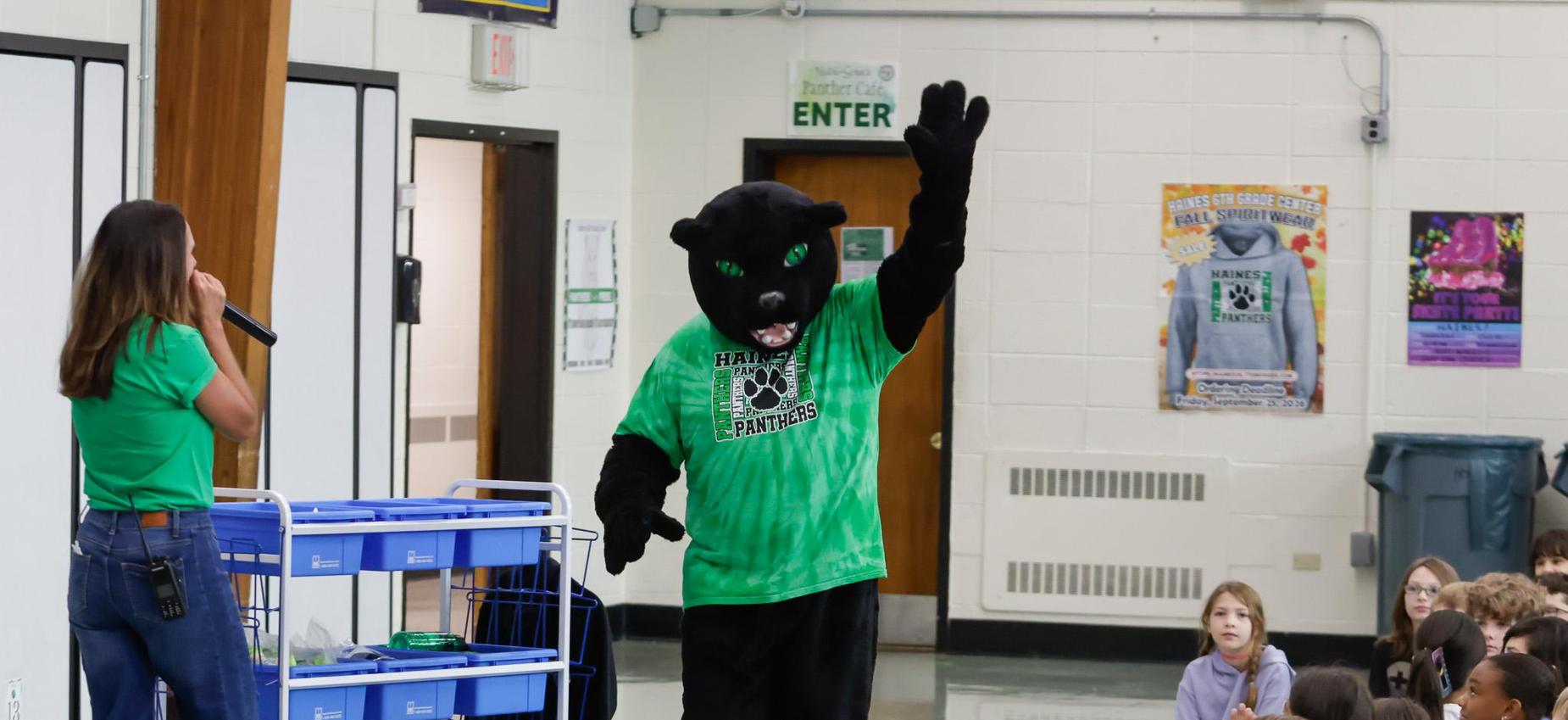 A person dressed in a panther mascot costume waves to students in an assembly.
