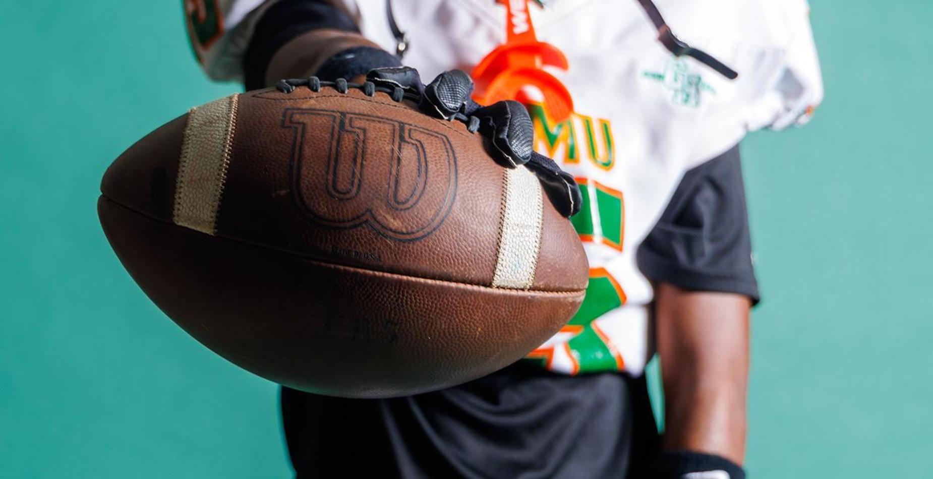 A player in a football uniform holding a football against a teal background.
