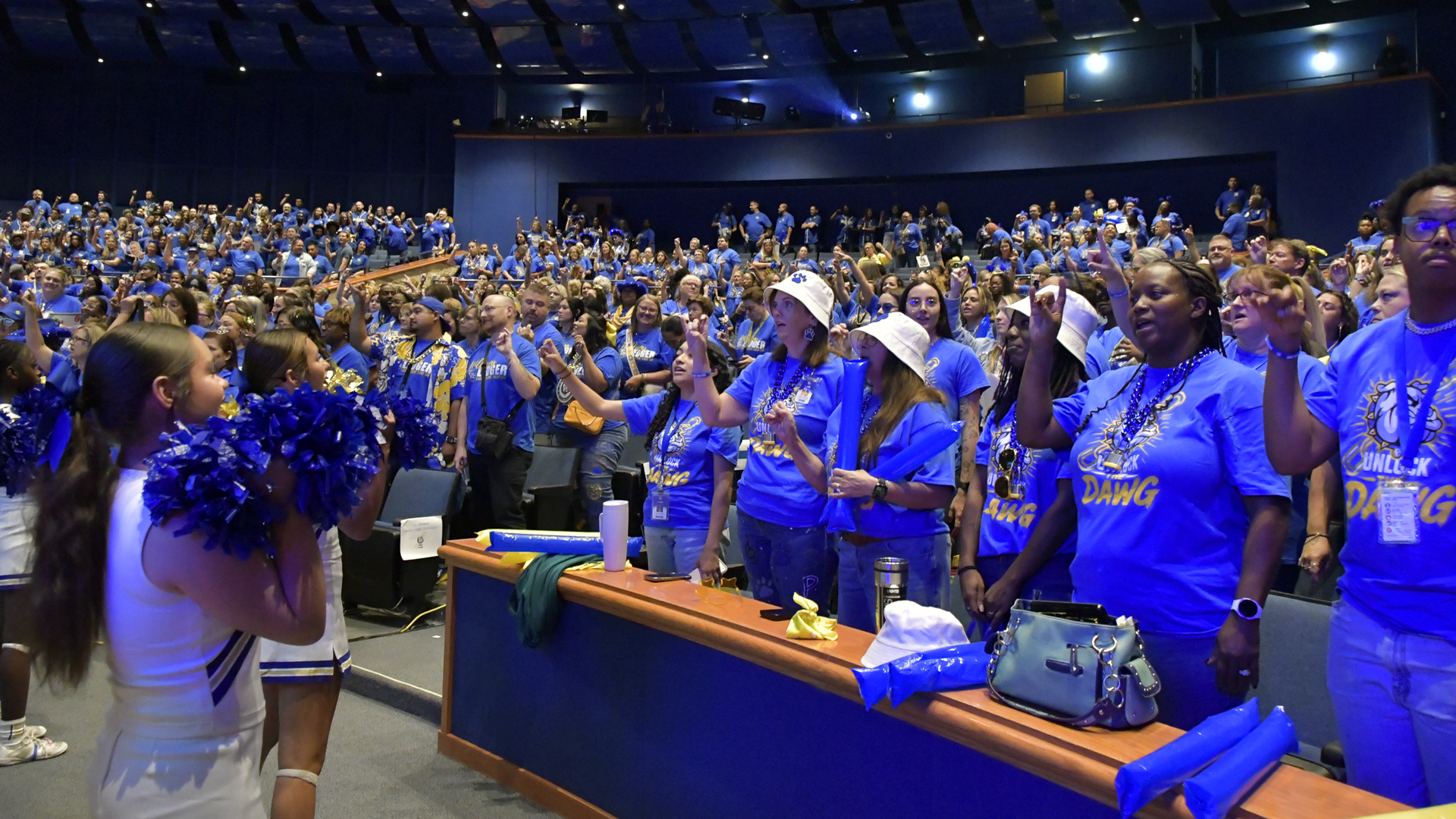 A large crowd of people in blue shirts at a lively event, with cheerleaders in front.