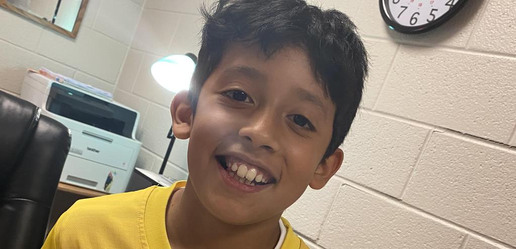 Boy in a yellow shirt smiles at the camera in a classroom.
