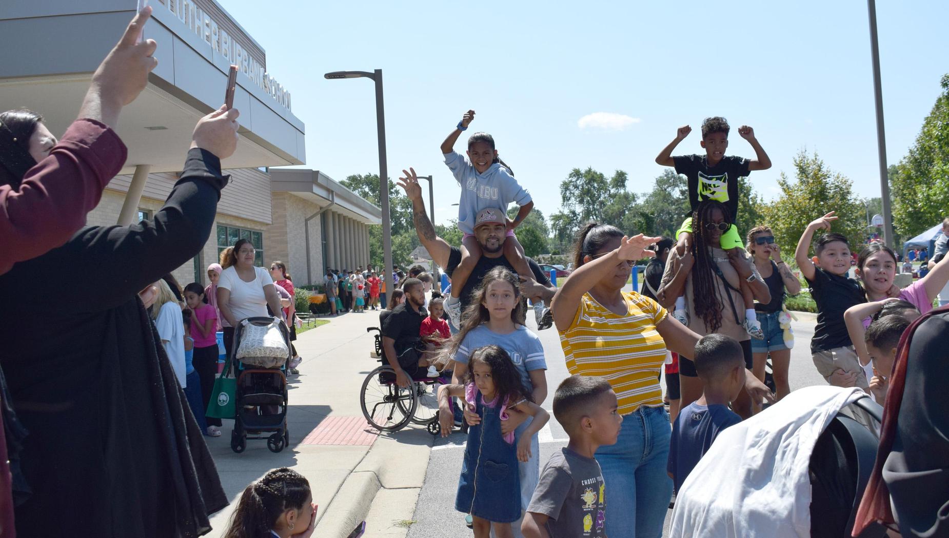 Crowd celebrating outside a school, with children on shoulders and people taking photos.