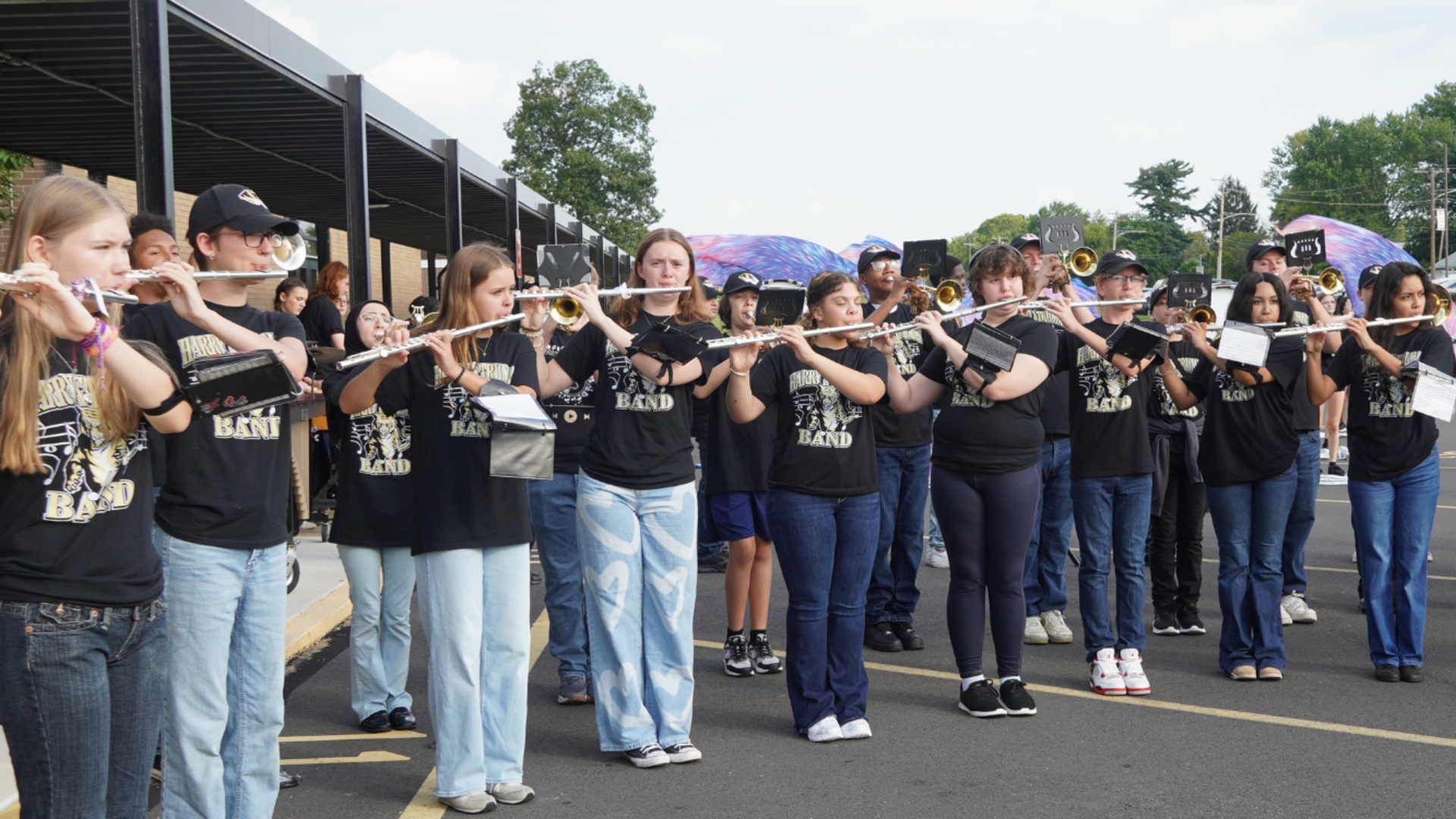 A large group of student marching band members in matching black t-shirts and jeans play flutes and brass instruments outdoors in the parking lot.