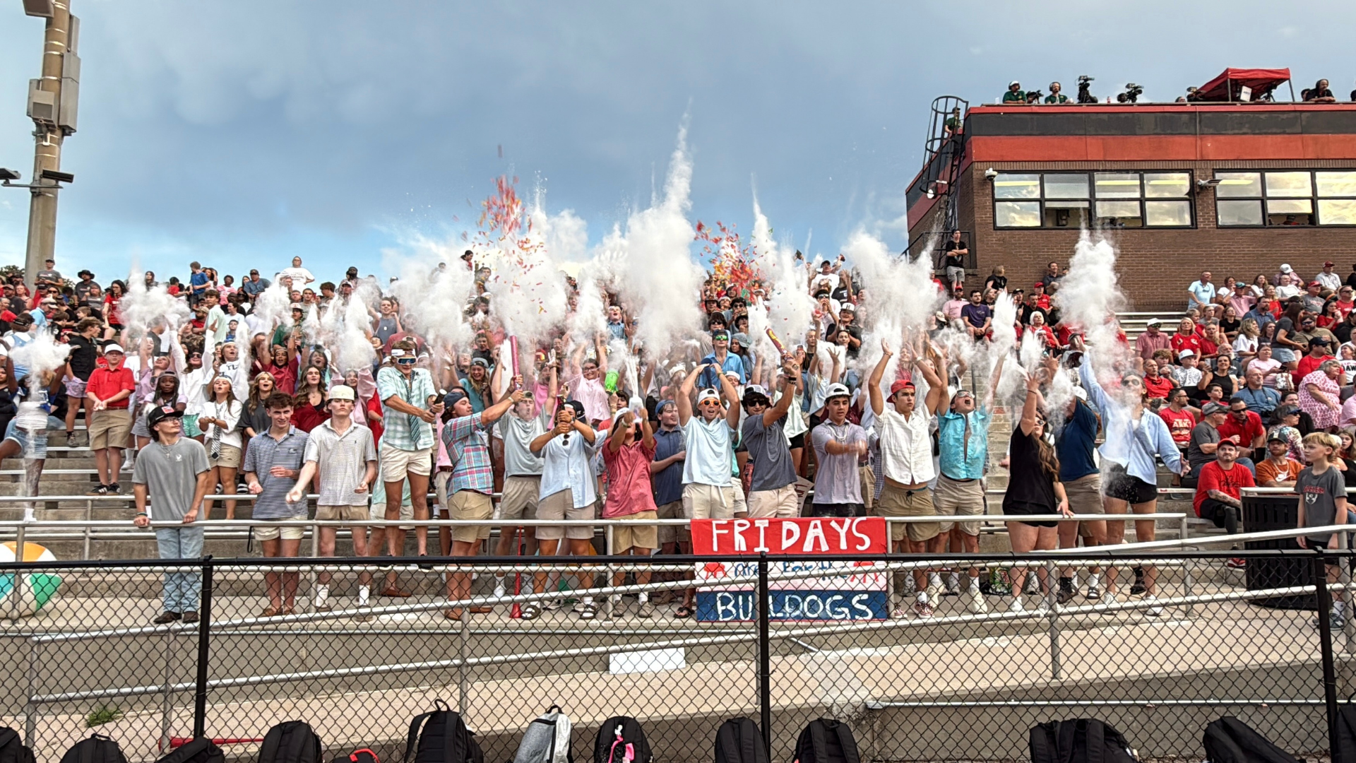 Cheering crowd at a sporting event throwing powder in the air with a sign that says 'FRIDAYS BULLDOGS'.