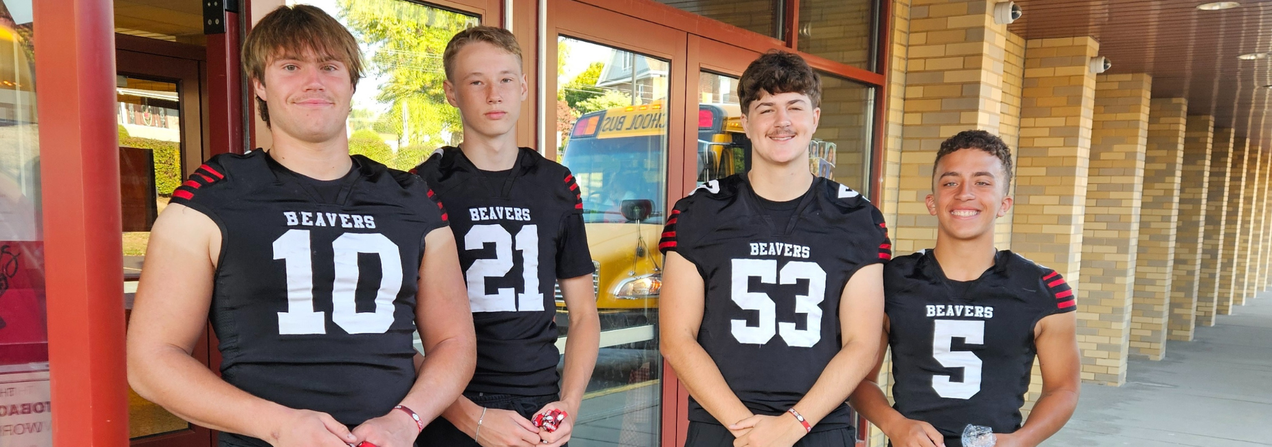 Four football players in black uniforms smiling outside a building with a school bus in the background.