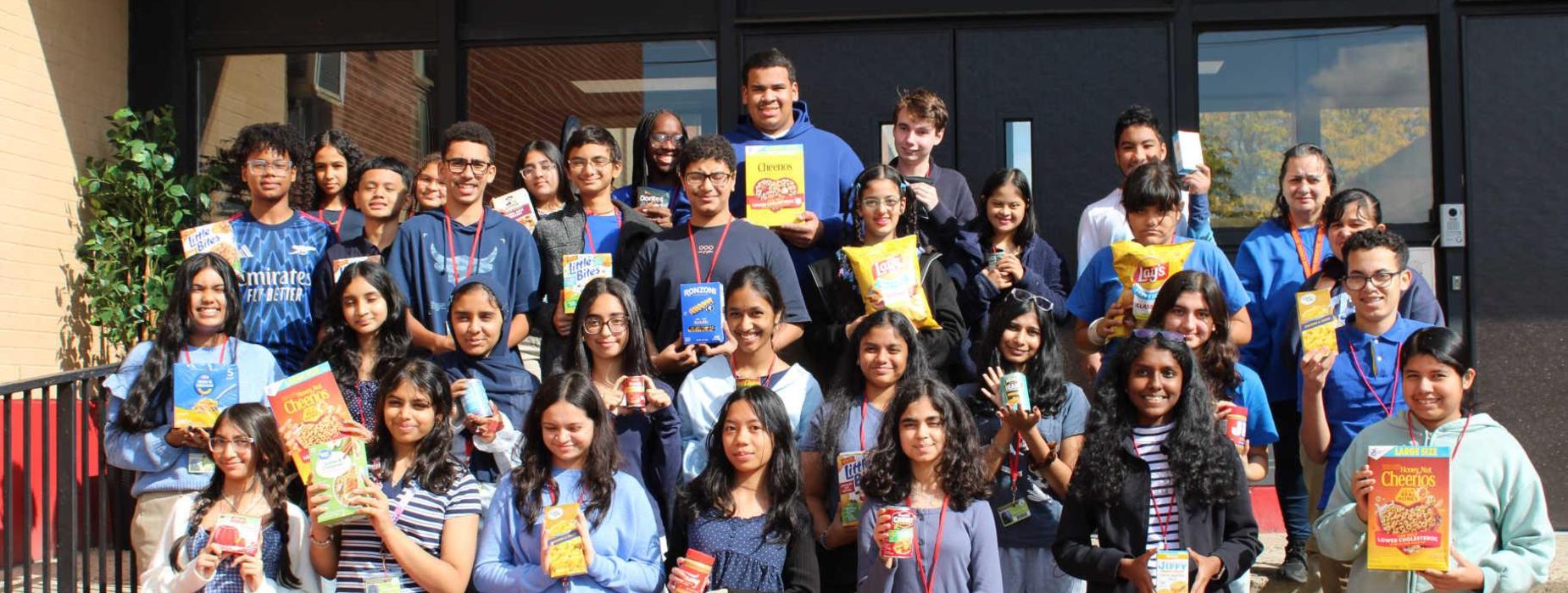 Group of students in blue shirts holding snacks, posing at a school entrance.