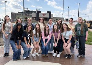A group of 15 young women poses outdoors with a school building in the background.