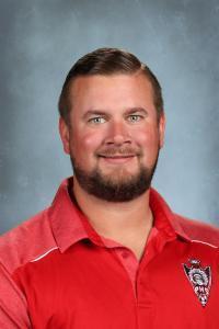 Smiling male teacher in a red shirt with a beard against a gray background.