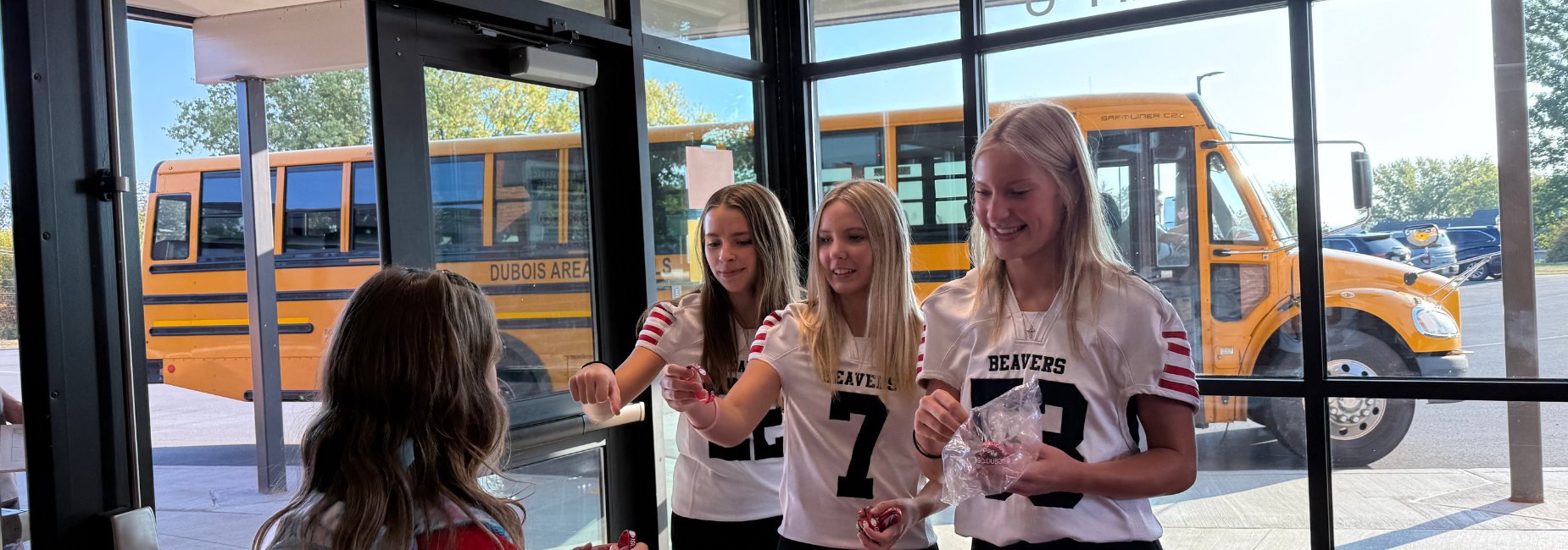 Three girls in jerseys handing out items at a school entrance with a bus in the background.