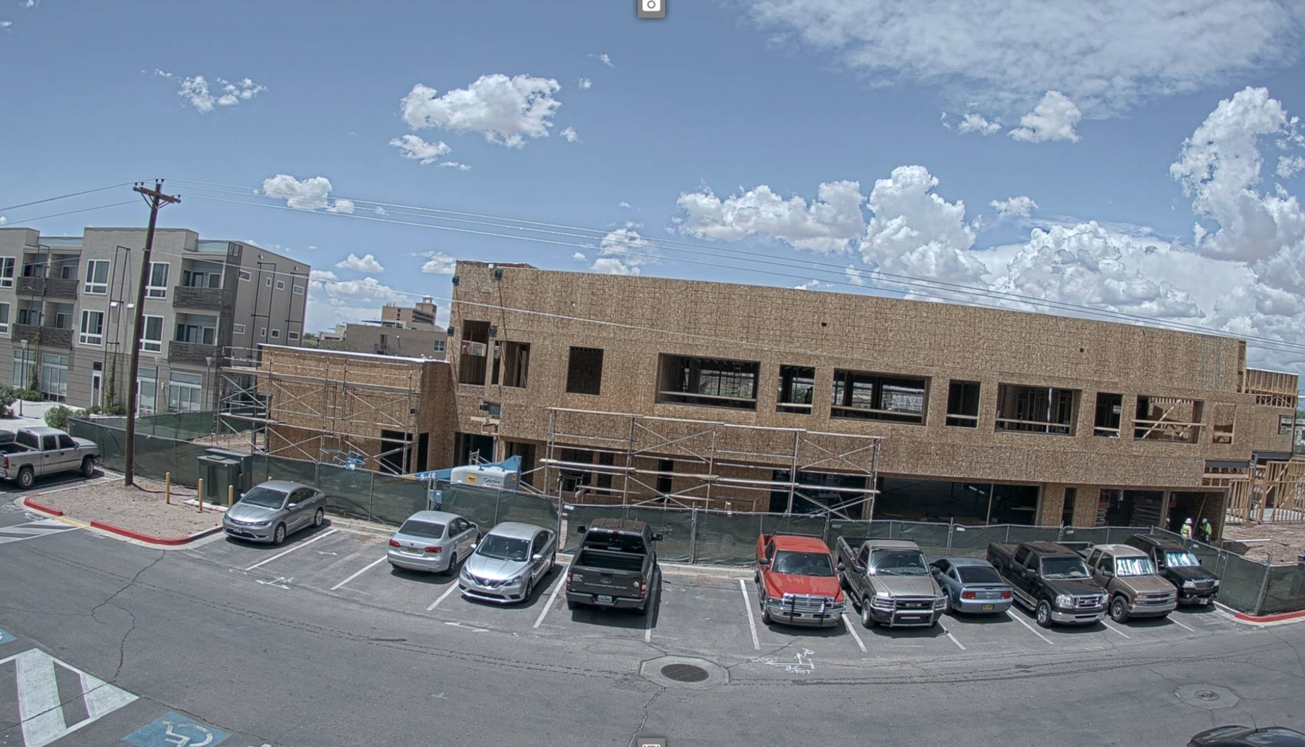 A photo of a building under a blue sky with some clouds. The building is under construction and the walls and framing are visible. Trucks and cars are parked in front of the building.