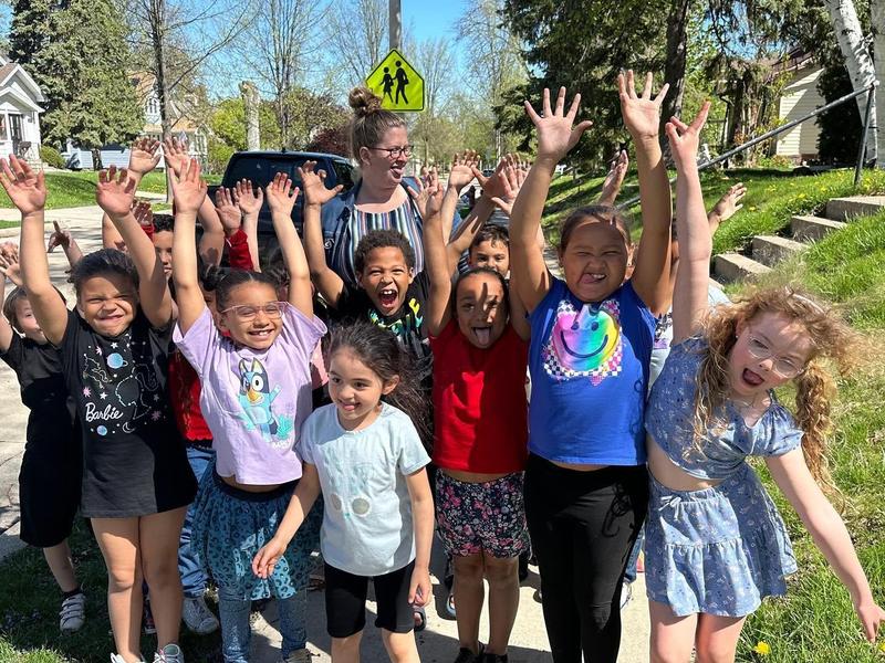 A group of children and an adult posing joyfully with their arms raised outdoors.
