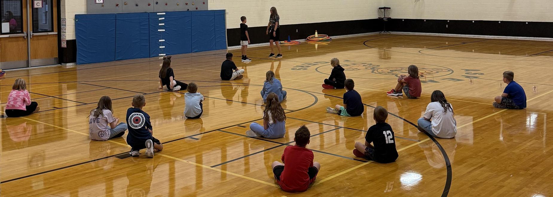 Students sitting in a gym during a class activity on the floor.