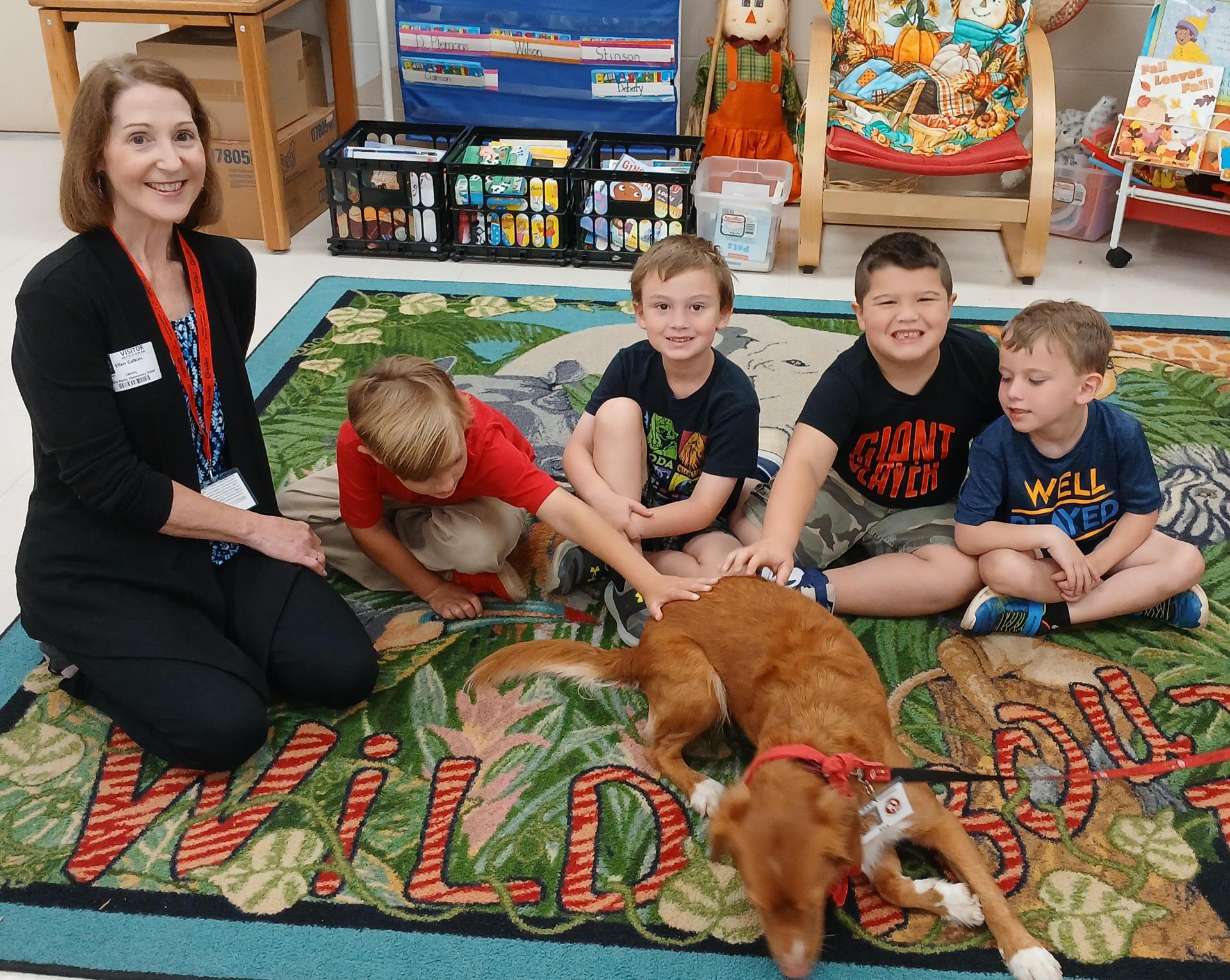 Students sitting on a colorful carpet in the library with a dog 
