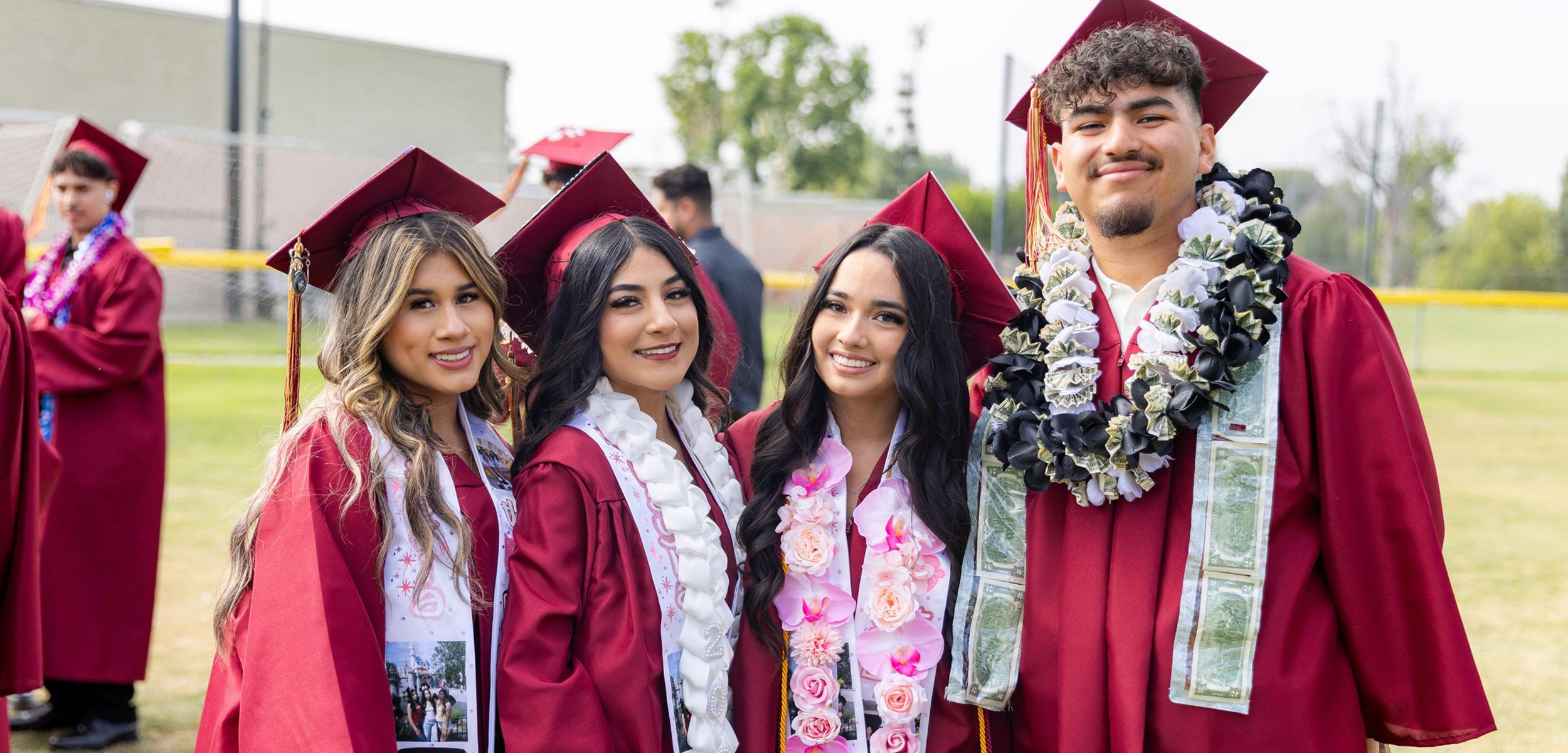 Four graduates in maroon caps and gowns pose together outdoors, smiling.