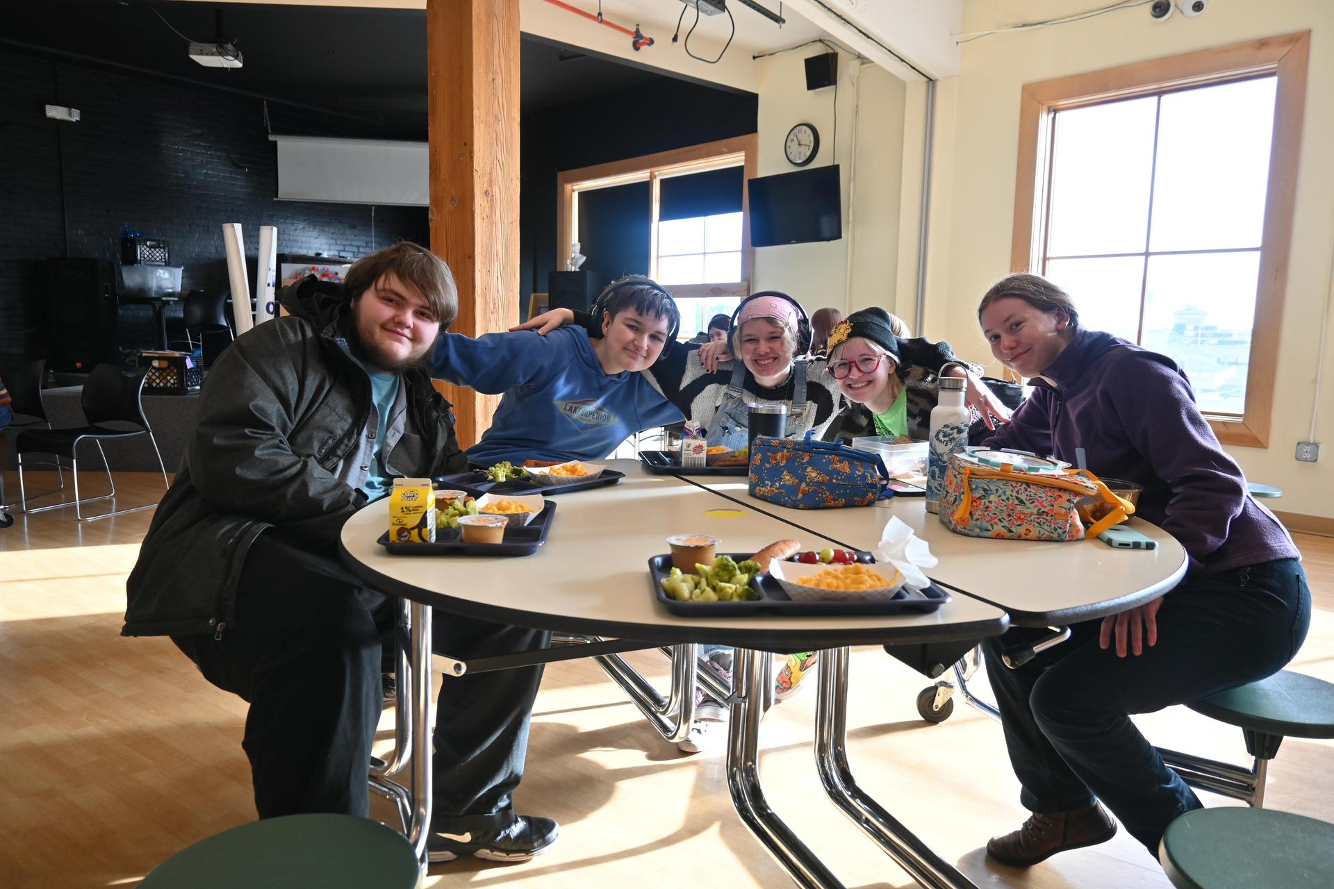 A group of 5 students gathered around a circular table, arms around each others shoulders, smiling.