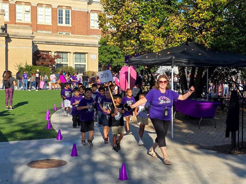 Children in purple shirts walking in a line with an adult in front, during an outdoor event.