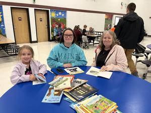 Two women and a girl seated at a blue table with various books in a school environment.