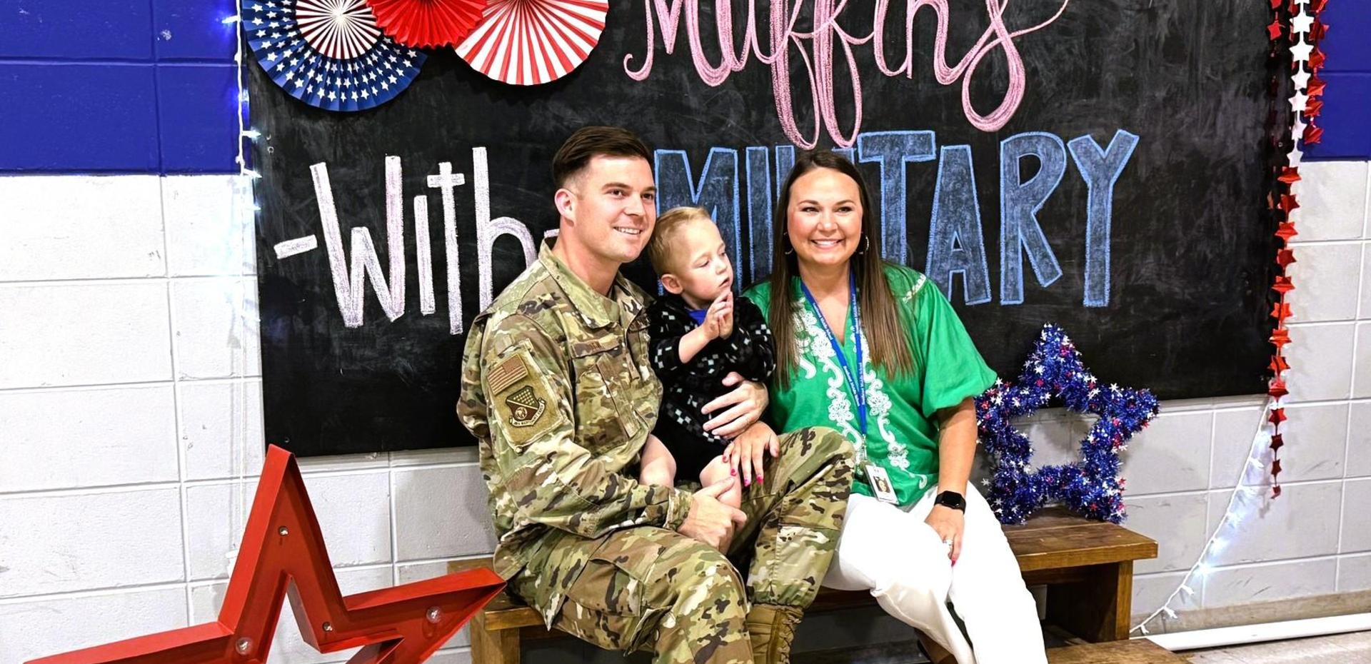 A soldier in uniform sits with a woman and a young child in front of a decorated backdrop.