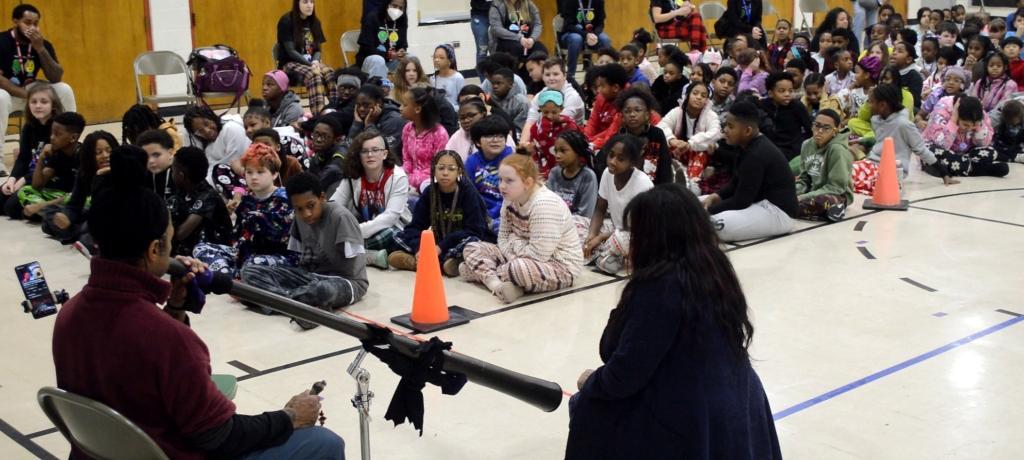 students sit on gym floor as man plays the didgeridoo