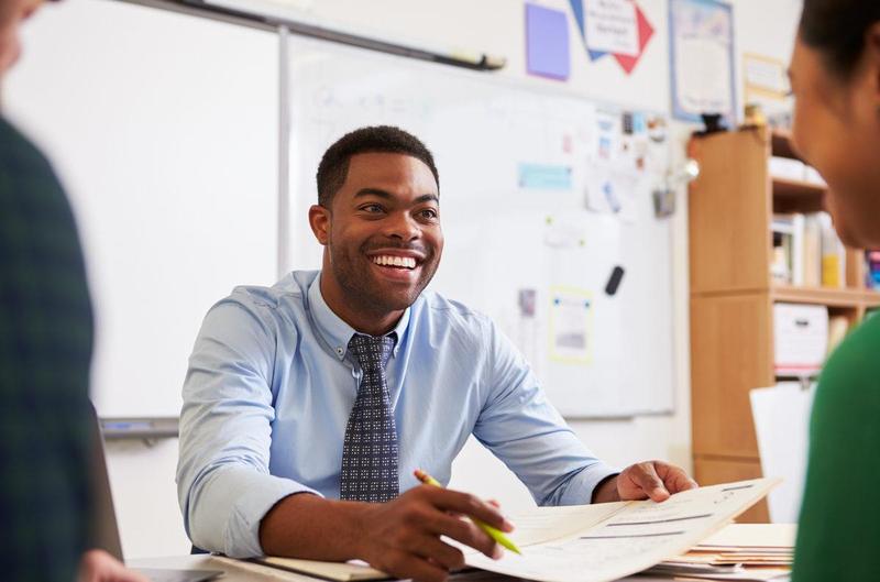 Smiling man in a blue shirt with a tie, discussing paperwork in a classroom.