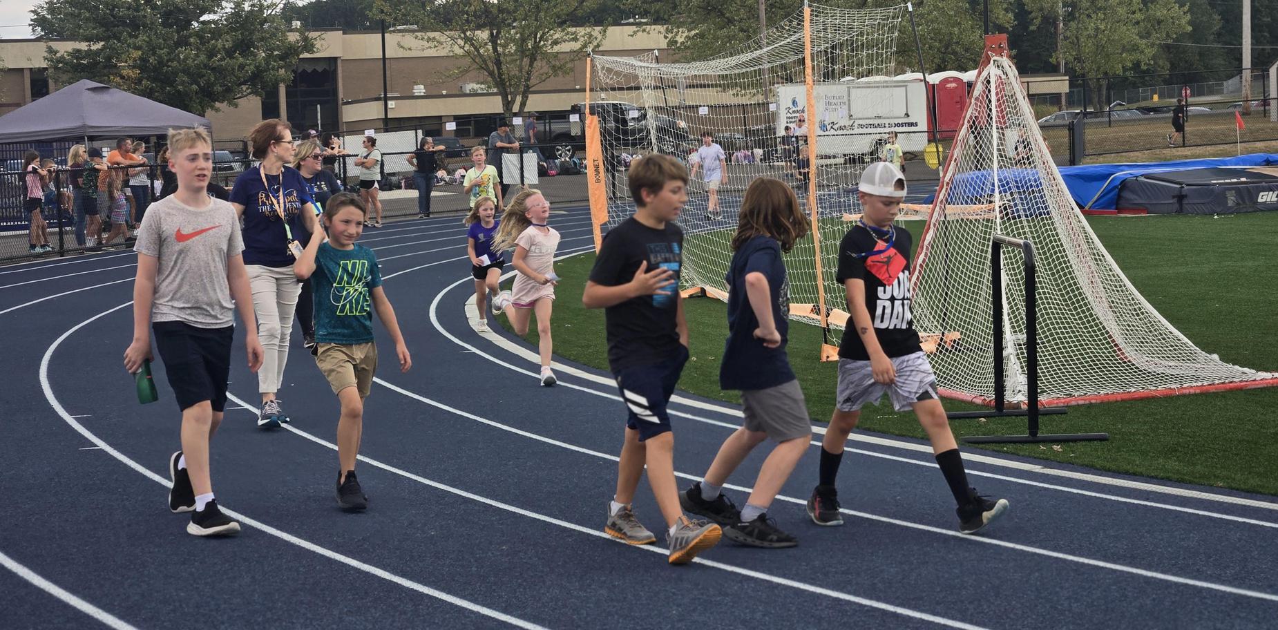 Group of children walking on a track, with parents and tents visible nearby.