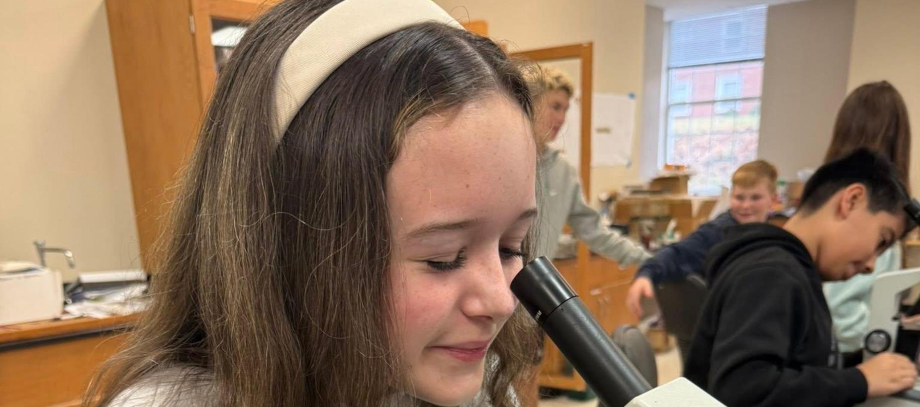 A student examines a slide under a microscope in a classroom setting.