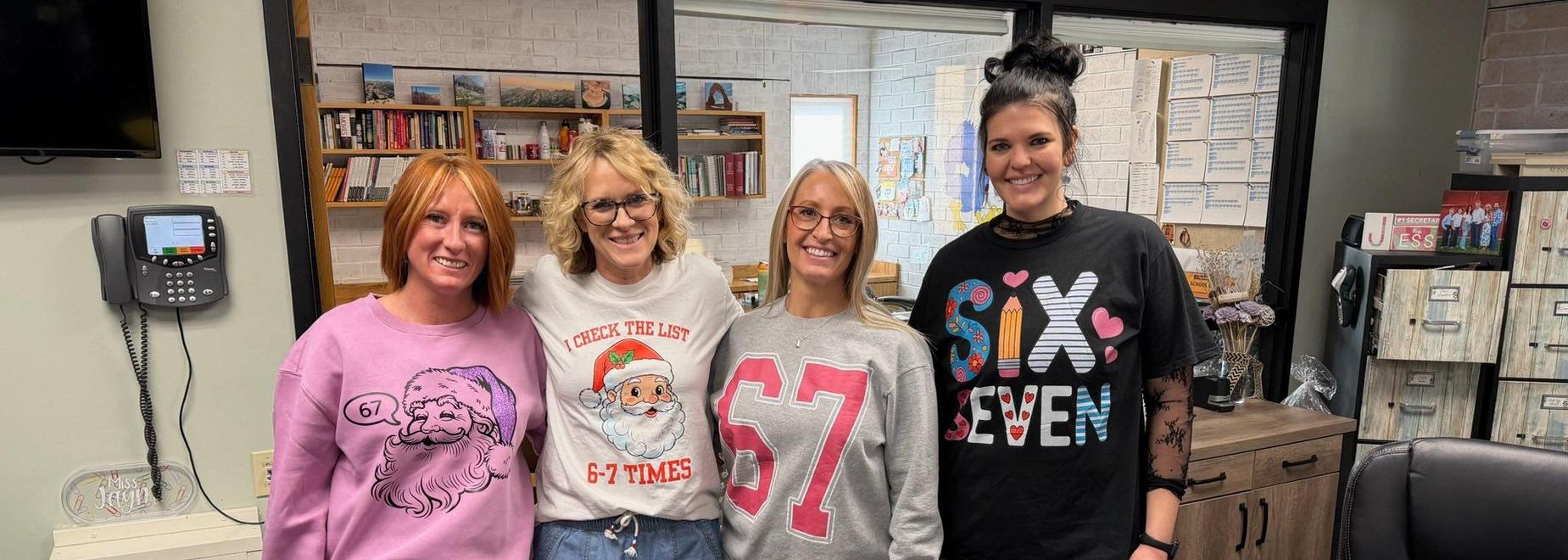 Four women wearing festive sweatshirts and smiling together in an office setting.