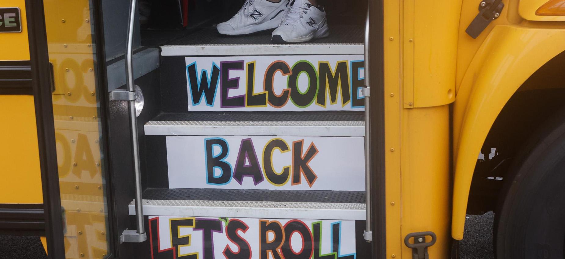 Colorful signs on school bus steps reading 'Welcome Back' and 'Let's Roll'