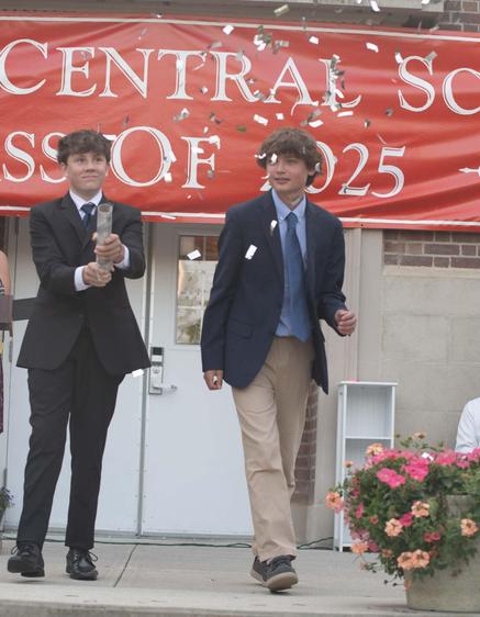 Two graduates walk celebrating under confetti in front of a school backdrop.