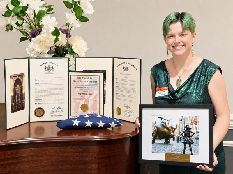 Image of a girl holding her drawing standing next to a table with her awards