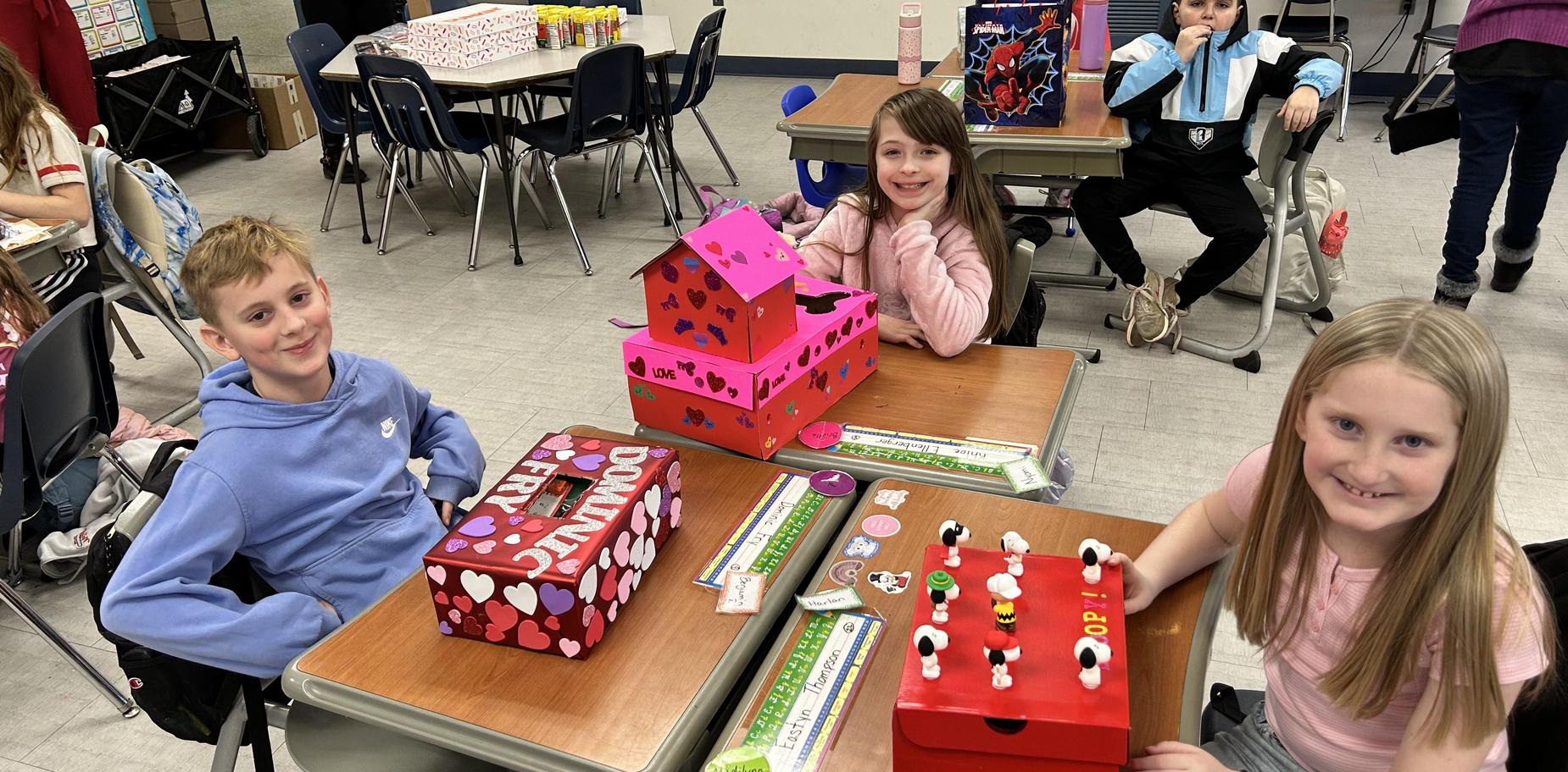 three students sit at desks with valentines boxes in front of them smiling