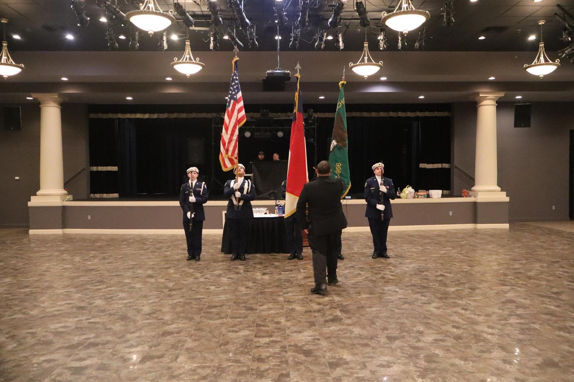 Four individuals in uniform presenting flags on a stage with seating in the background.