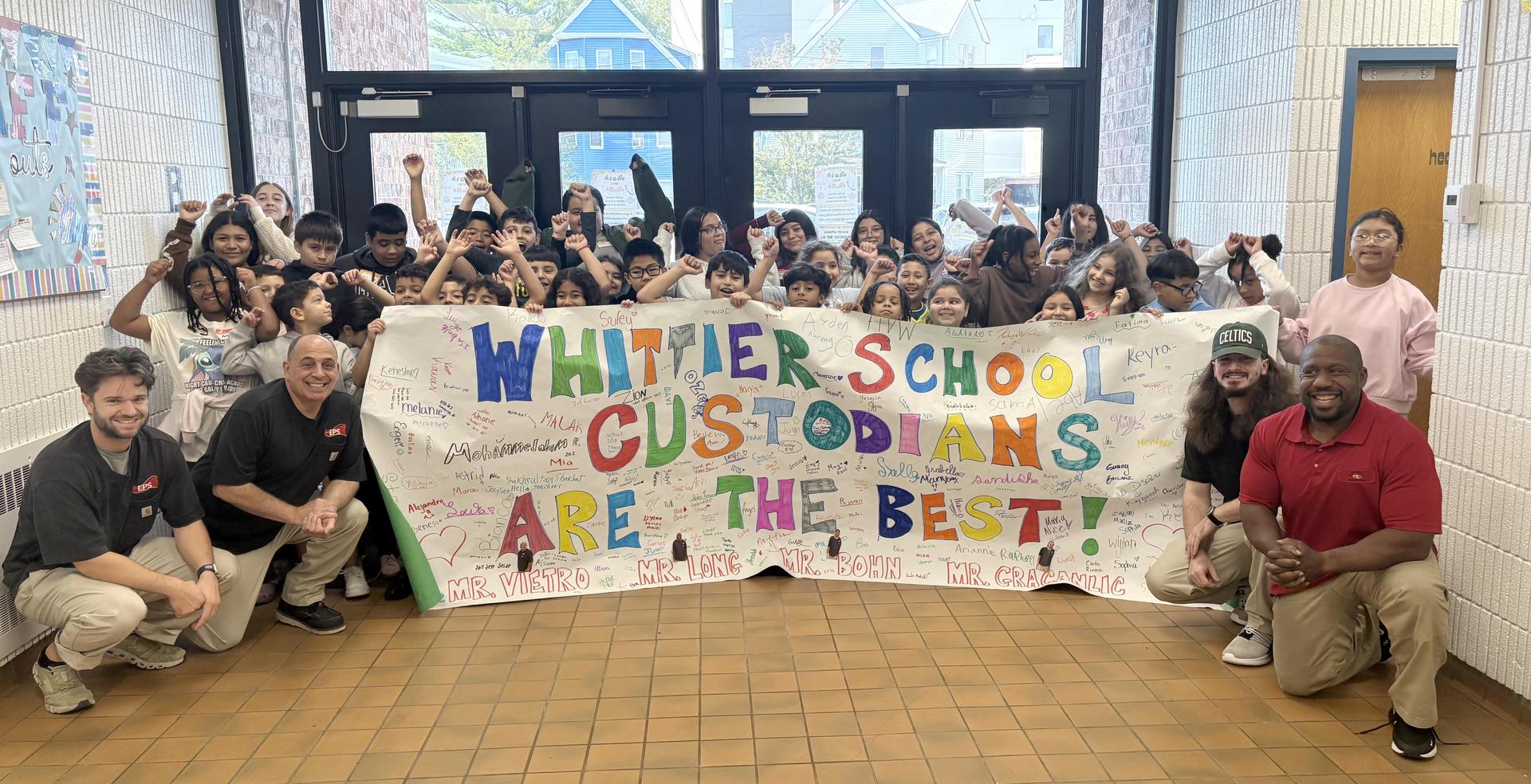 Students and staff celebrate with a banner praising custodians at Whittier School.