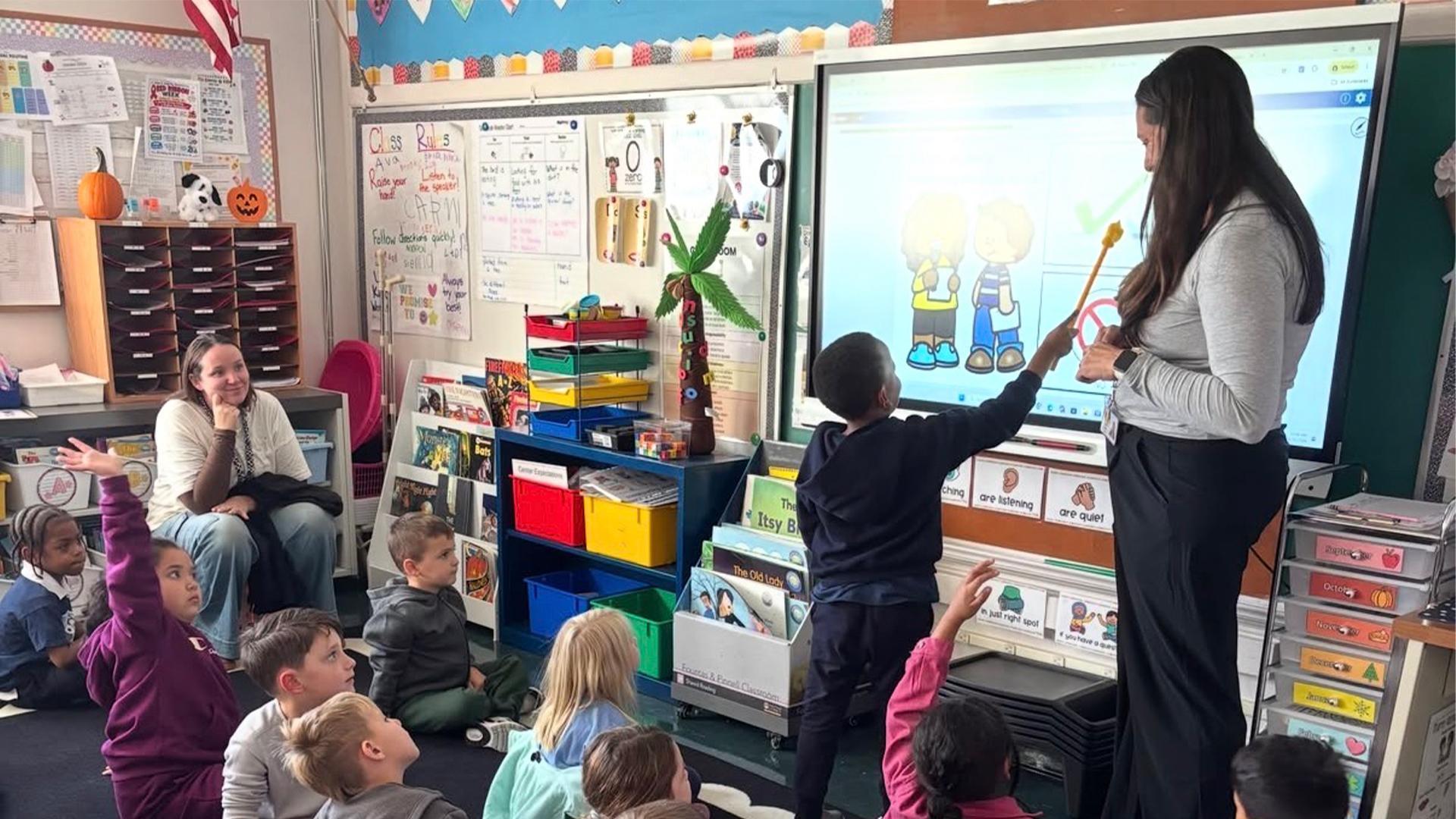 Classroom scene with students engaged in a lesson and a teacher using a digital display.