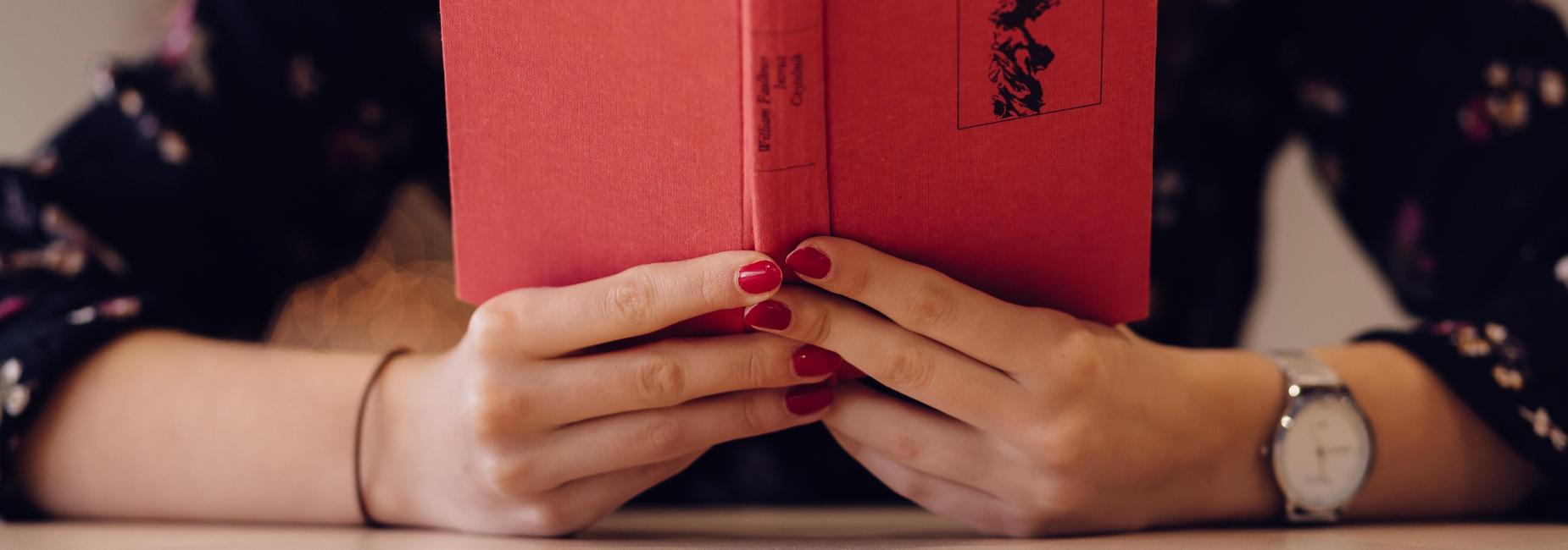 A person holding a red book with floral patterns on the cover and painted nails.